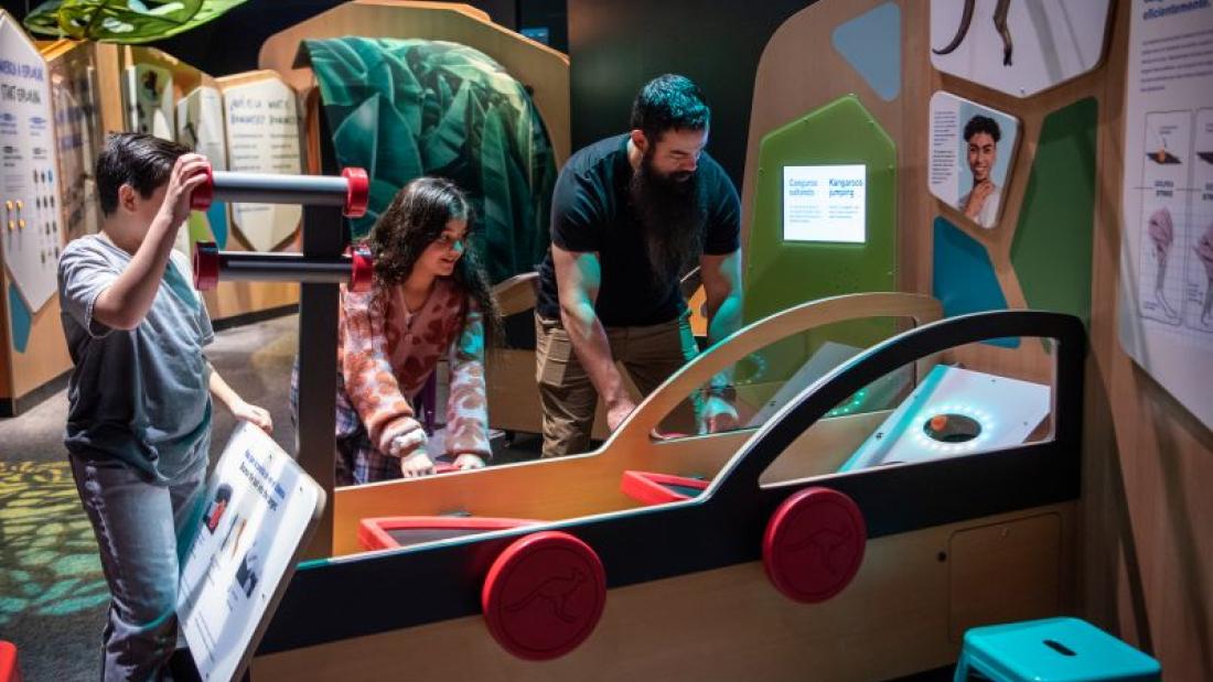 A boy watches a girl and man interacting with an exhibit piece shaped like a car