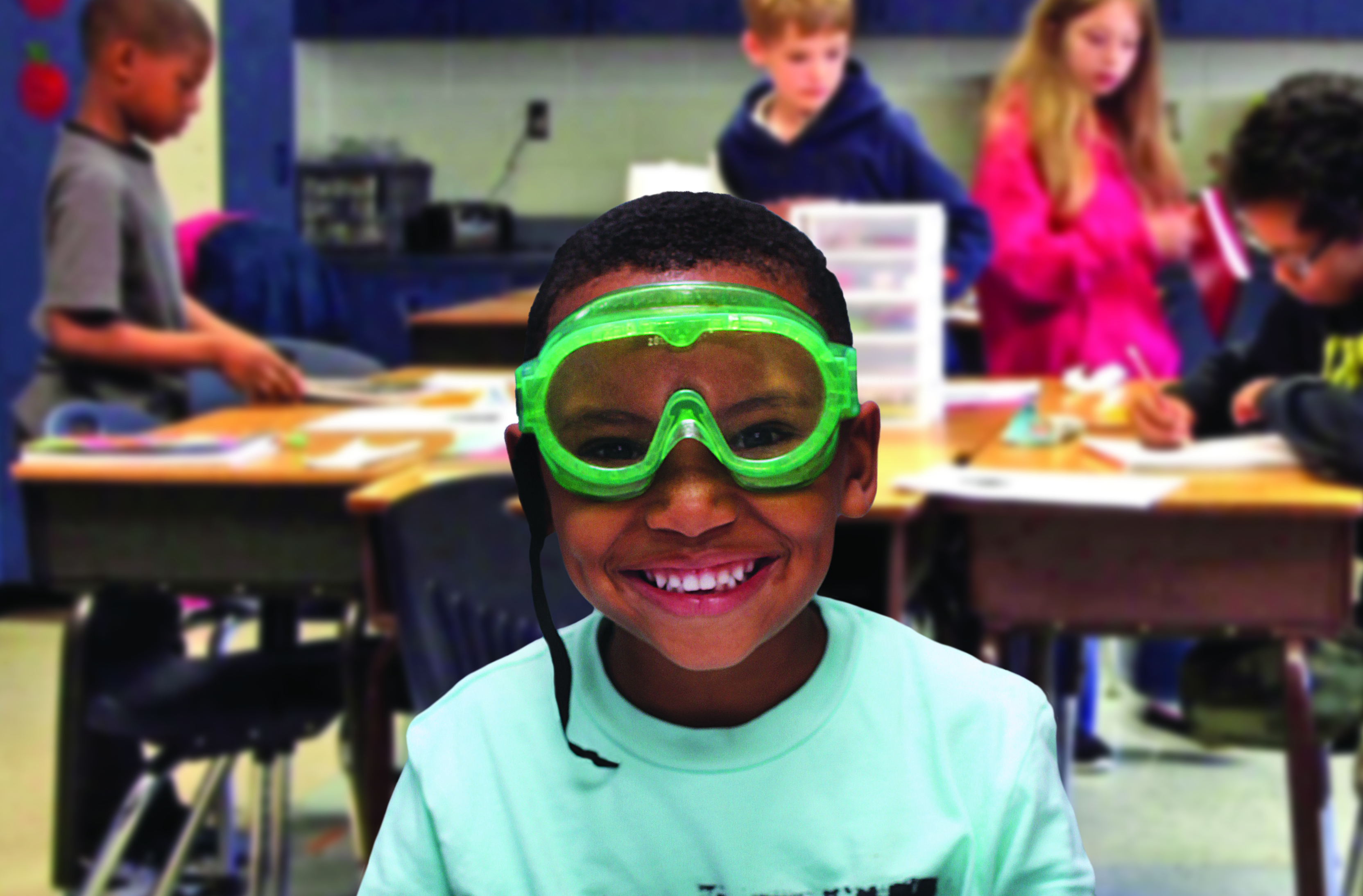 A young black boy wearing green safety goggles in a blue shirt smiles at the camera in a classroom