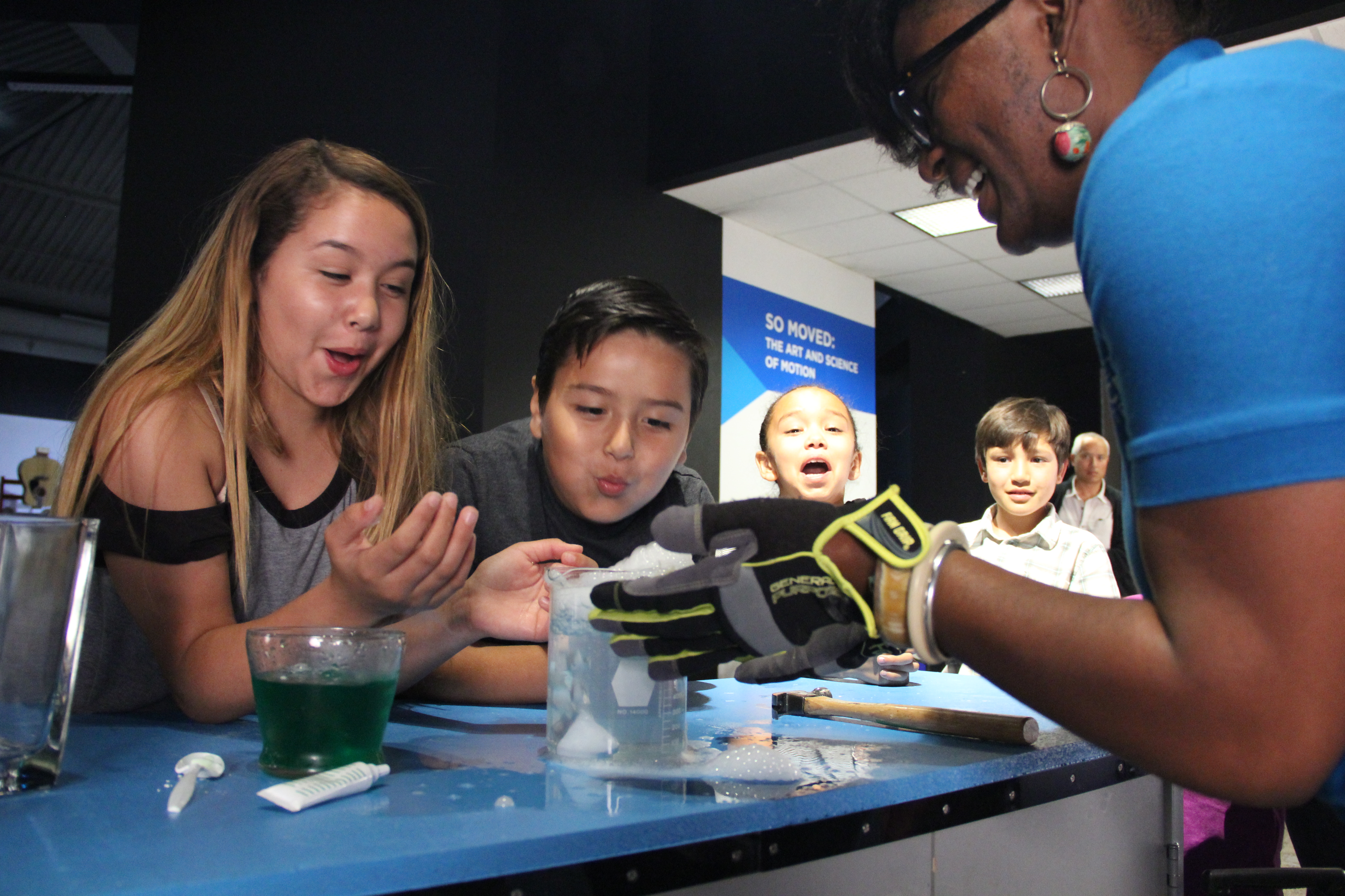 Four young hispanic kids, three girls and one boy, lean over an experiment with excitement while a black woman wearing safety gloves holds a foaming science beaker