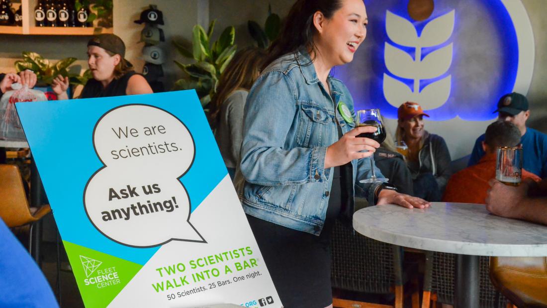 A sign in the foreground says We Are Scientists Ask Us Anything. A woman in the background holds a drink and talks with a smile