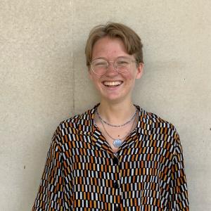 A white person whose pronouns are They/Them. They have short cropped blonde hair, silver glasses, two silver necklaces, and is wearing a loose fitting buttonup top that has a orange, yellow, and white repeating pattern. They are standing against a tan concrete wall. 