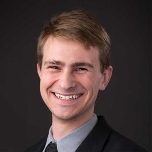 A white man with short blonde hair smiles in a formal headshot. He is wearing a black suit jacket, dark tie, and grey collared shirt. The background is a photographers neutral grey.  