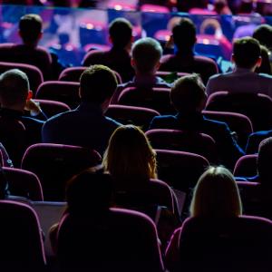 A view of people sitting in theater style chairs in a darkened room from behind. The people are front lit so we only see the outlines of the seated people. The top of the image is brighter and has a purple and blue glow.