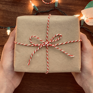 Brown paper wraped gift with a red and white stripped bow being held by a pair of hands with a table and holiday lights in the background.