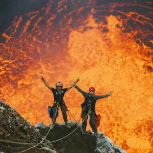 Two people tethered to ropes have repelled down into a volcano and are hanging above a pt of lava