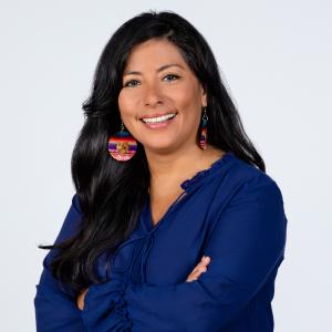 A woman with long dark hair tucked behind one ear and one shoulder wearing a blue blouse with multicolored crafted round earrings. She is smiling and standing with arms crossed in front of a neutral background