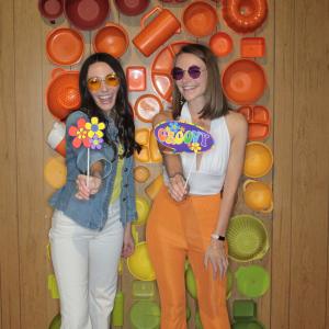 Two young women pose in front of a bright wall display while wearing 70s inspired clothes and holding flowers signs and a groovy sign 