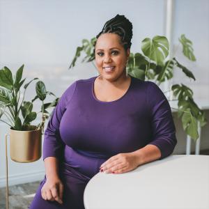 A black woman in a purple shirt sitting at a white table with green plants in the background