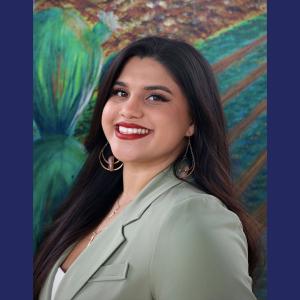 A hispanic woman in a light green business jacket smiling against a painted mural background 