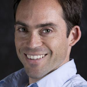 Professor Adam Burgasser. Portrait of a man on a dark background with a blue shir smiling.t