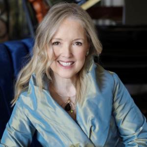 Dr. Julie Cowan Novak. A woman with long, wavy blond hair smiles warmly at the camera. She is wearing a light blue, silky jacket, and a necklace featuring a large black pendant with a green accent. The background includes a hint of musical instruments, suggesting a creative or artistic environment.