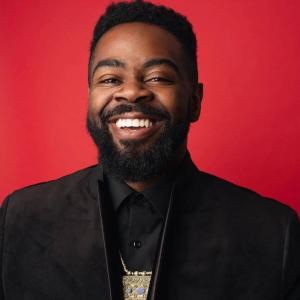 The image captures a man with a warm and inviting smile set against a striking red backdrop, a color choice that amplifies the photo's vibrancy and draws immediate attention to the subject. The man has dark hair and a neatly trimmed beard, which contributes to his well-groomed appearance. His smile is wide, revealing a full set of teeth that add to his approachable and friendly demeanor.  He is dressed in a sophisticated black suit, which contrasts with the bright background and gives the photo a classic an