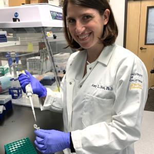 Amidst a laboratory backdrop, a woman scientist stands confidently, dressed in a crisp white lab coat. The lab coat features her name, Amy L. Non, Ph.D., elegantly embroidered on the left side, while the seal of the University of California adorns the right sleeve.